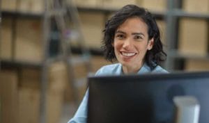 Smiling person with dark hair in a light blue shirt sits at a desk, managing supply chain tasks on a computer monitor. Blurred background features shelves with boxes.