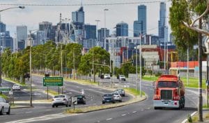 City road with cars and truck driving in different directions. Tall skyscrapers are in background, with trees lining the road.