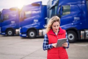 A woman wearing a red vest is using a tablet in front of a row of blue trucks parked in a lot. She's focused on mastering freight management.