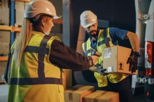 Two warehouse workers wearing hard hats and high-visibility vests are handling a box. One worker is scanning the label on the box with a handheld scanner, while the other is holding it, ensuring accurate freight tracking. They are surrounded by more boxes and storage shelves in the background.