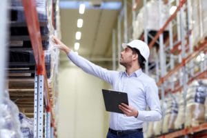 Person with white hard hat and light blue shirt is involved in freight management, holding a clipboard and reaching for items on a high warehouse shelf. With tall shelves filled with various boxed goods in background.