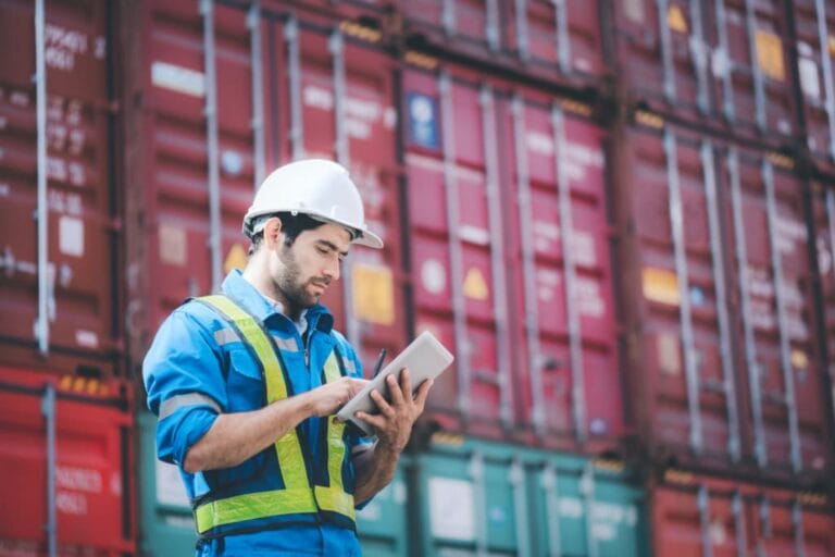 A construction worker in a white hard hat and reflective vest uses a tablet, expertly navigating shipping management tasks in front of stacked red and green containers. The worker appears focused on optimizing the logistics displayed on the device.