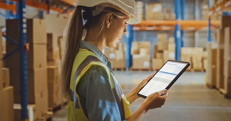 A person wearing a hard hat and a safety vest stands in a brightly lit, organized warehouse, holding a tablet. They are looking at the screen, reviewing freight paperwork, with shelves stocked with boxes and various items in the background.