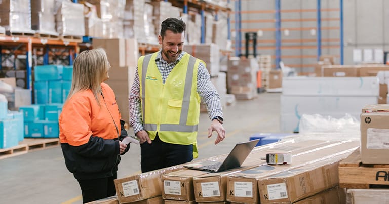 A man and a woman in a warehouse engage in a conversation about sustainable shipping. The man, wearing a Macship yellow safety vest, points at a laptop on some stacked boxes, while the woman, in an orange safety jacket, listens attentively. Background shows is in a warehouse with racks and stocks