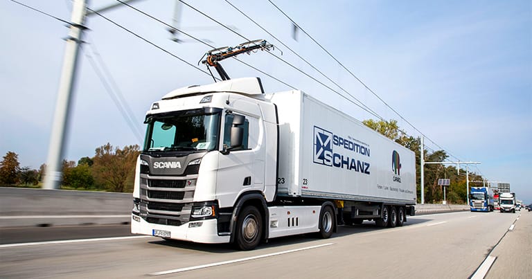 A white semi-truck with "Spedition Schanz" branding is driving on a highway, connected to overhead electric power lines, suggesting it's an electric or hybrid vehicle. This highlights the company's commitment to sustainable shipping. Trees and another truck are visible in the background.