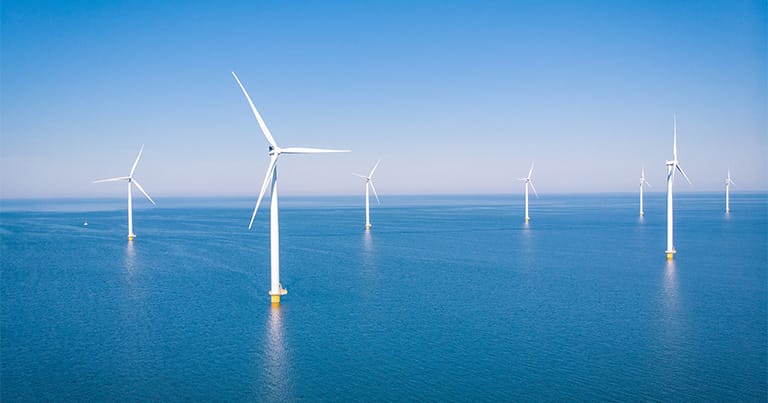 Offshore wind farm with multiple white turbines standing in the calm blue ocean under a clear sky. The turbines are evenly spaced, harnessing wind energy to generate electricity. In harmony with sustainable shipping efforts, the far horizon blends seamlessly with the sky.