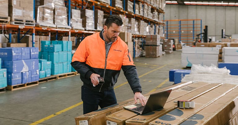 A warehouse worker in a bright orange and navy jacket uses a laptop on a workbench surrounded by large boxes and packages. Shelves filled with more boxes and products are visible in the background. The worker appears focused, holding a barcode scanner, likely optimizing freight management services.