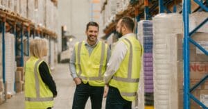 Three people wearing Machship yellow safety vests are standing and discussing the latest freight news in a warehouse aisle. The shelves around them are stocked with various goods.