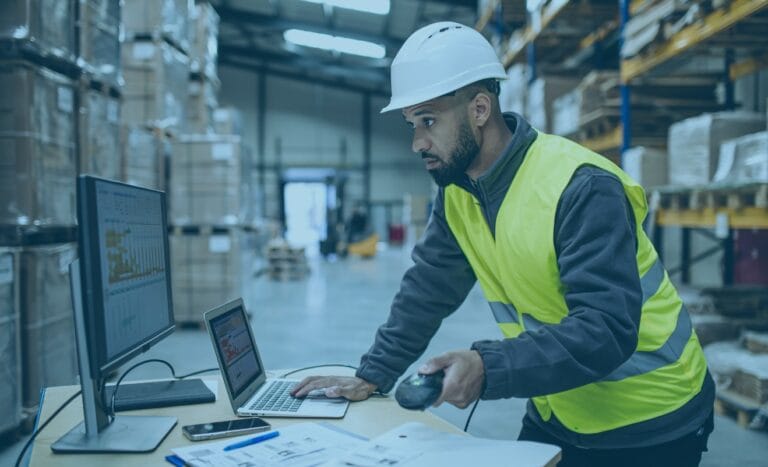 Man in safety gear using a laptop and desktop for freight management system in warehouse.