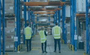 Three freight leaders in yellow vests walk through a warehouse with shelves of supplies.