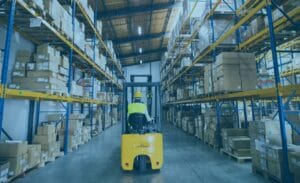 Worker drives a yellow forklift, boosting supply chain efficiency in a box-filled warehouse aisle.