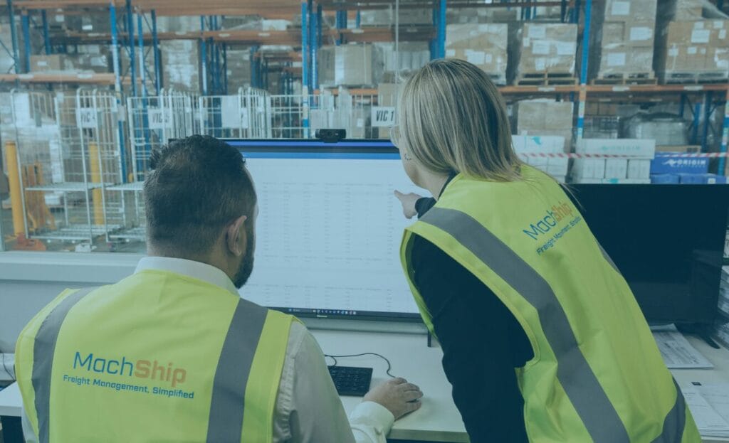 Two people in safety vests review data on a computer screen in a warehouse office.