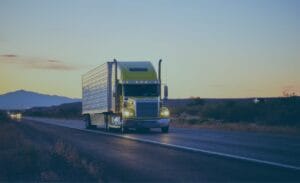 A green semi-truck driving on a highway at sunset with mountains in the background.