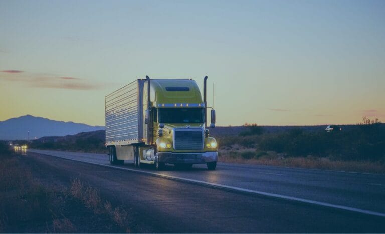 A green semi-truck driving on a highway at sunset with mountains in the background.
